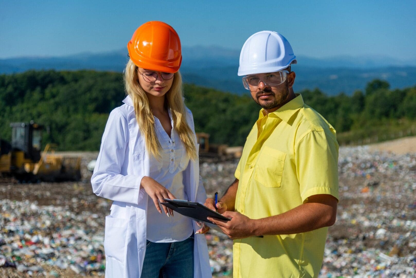 Two people wearing safety helmets and glasses stand at a landfill site. One holds a clipboard, and they both appear to be discussing something.