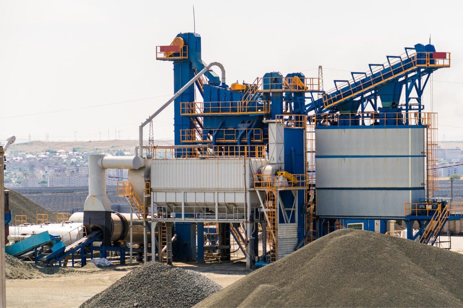 Industrial plant with blue and orange machinery for processing materials, surrounded by piles of gravel, set against a backdrop of a distant cityscape.