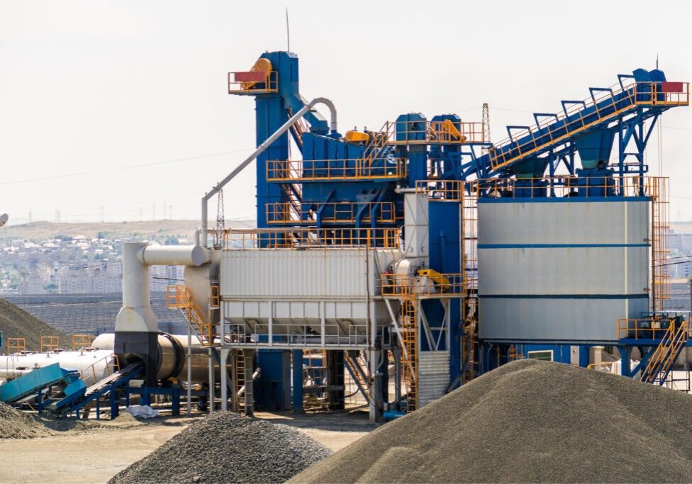 Industrial plant with blue and orange machinery for processing materials, surrounded by piles of gravel, set against a backdrop of a distant cityscape.